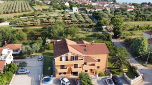 Un edificio de varios pisos con techo de tejas rojas, rodeado de vegetación y coches aparcados.