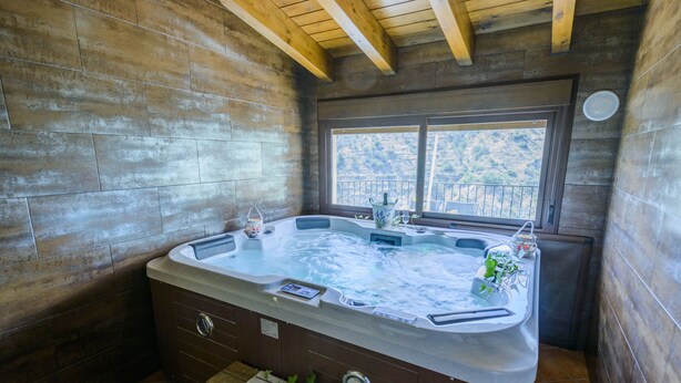 A hot tub in a rustic cabin with wooden walls, a window overlooking a mountainous landscape, and a small table with a plant. - RURAL HOUSE "TENGUERENGUE" (Laroya, Almería)