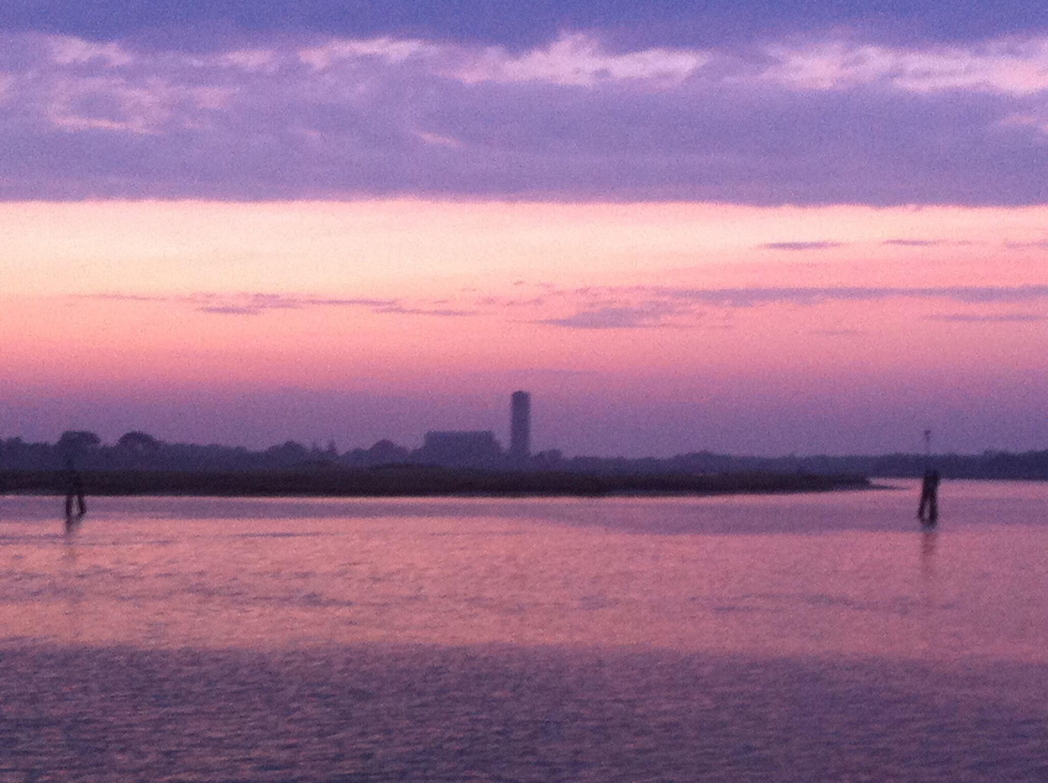 A sunset over a calm body of water with silhouettes of trees and a distant building.