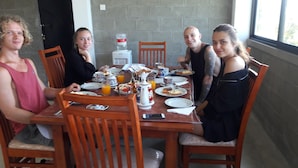 Un grupo de personas sentadas alrededor de una mesa de madera con desayuno, un dispensador de agua y una ventana al fondo.