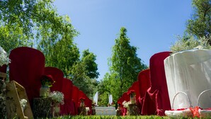 Une allée de cérémonie de mariage agrémentée de chaises rouges et de tables décorées de blanc, le tout entouré de verdure et d’arbres.