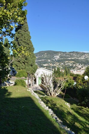 A lush garden with a tall tree, a small building, and a view of a town in the background.