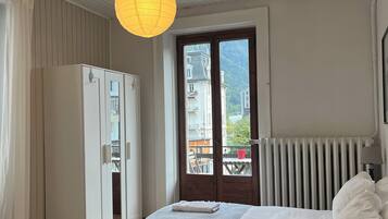 Une chambre à coucher comprenant un lit, une armoire, un radiateur et une vue sur des immeubles, visible à travers une porte.