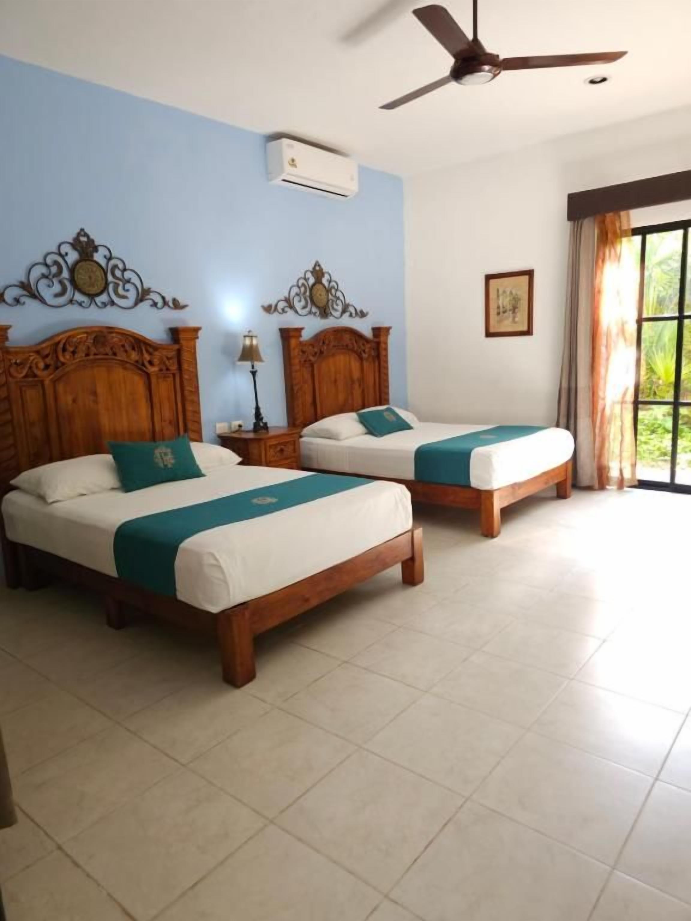 Habitación de hotel con dos camas de madera, un ventilador de techo y un gran ventanal con cortinas.