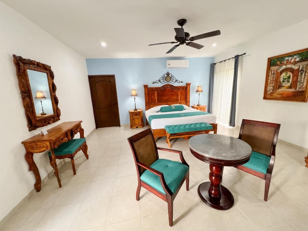 Una habitación de hotel con cama, dos sillas, una mesa, un ventilador de techo y un cuadro en la pared.