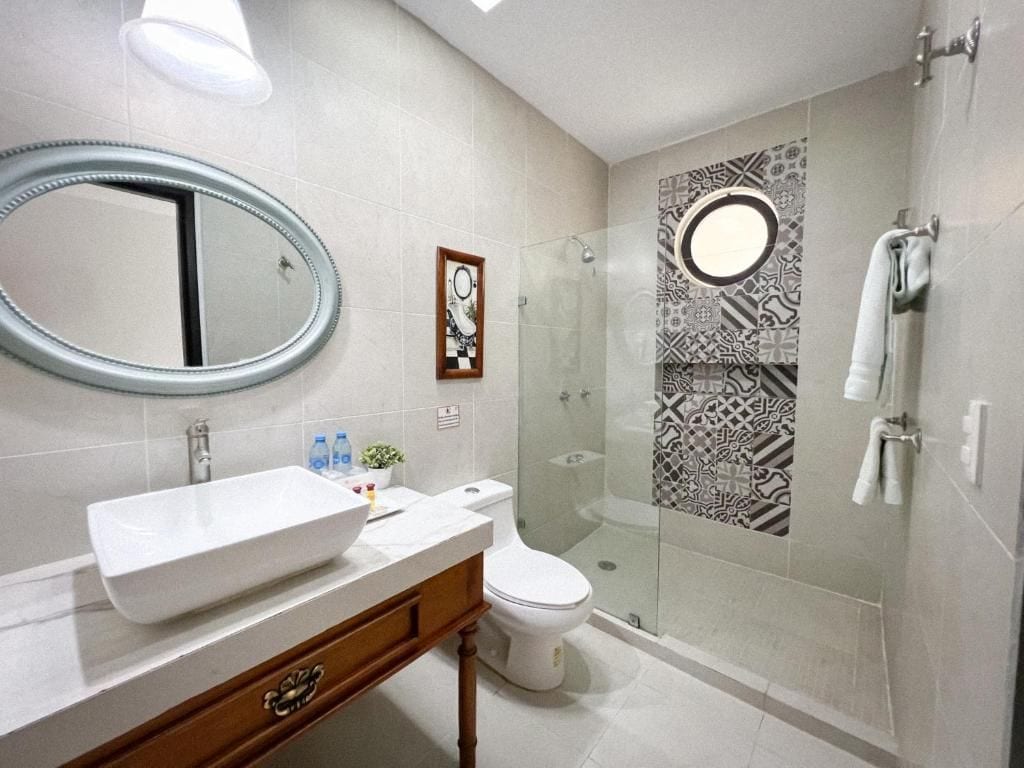 Un baño moderno con un espejo ovalado grande, un lavamanos blanco, un inodoro y una zona de ducha con mampara de cristal.