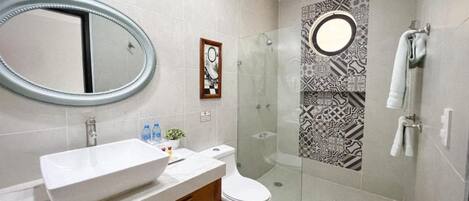 Un baño moderno con un espejo ovalado grande, un lavamanos blanco, un inodoro y una zona de ducha con mampara de cristal.
