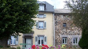 Un edificio de dos pisos con fachada de piedra y enlucido, un patio cubierto de césped y un cantero con flores rojas y púrpuras.