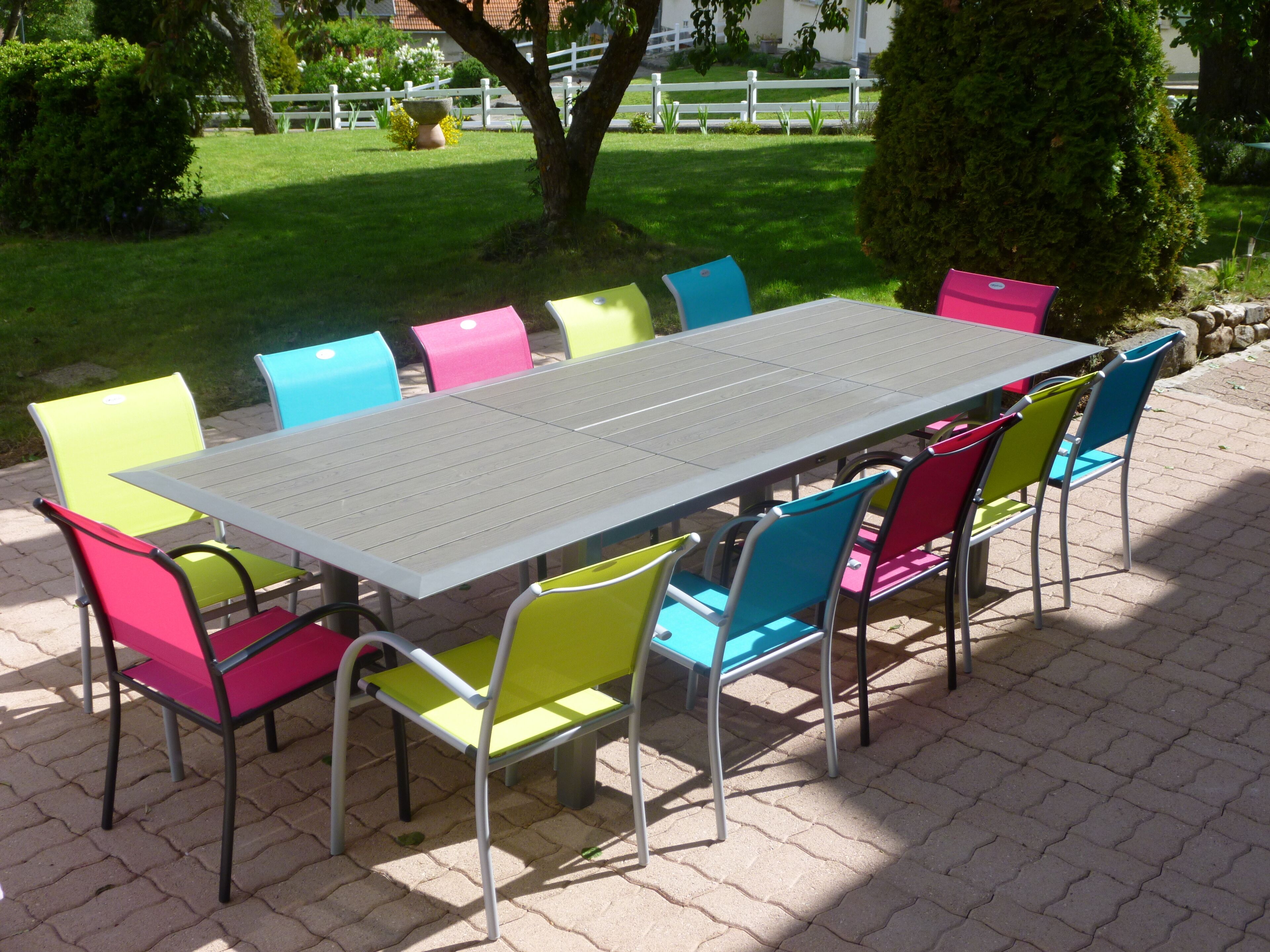 A rectangular outdoor table with a grey surface and metal legs, surrounded by colorful chairs with metal frames and cushioned seats in various colors.