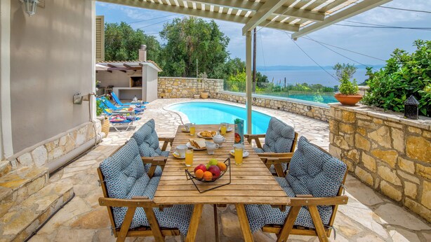 A covered outdoor seating area with a pool, stone walls, and a view of the sea.