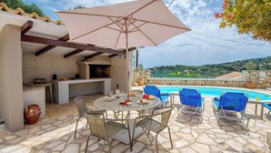 An outdoor patio with a stone floor, a round table with chairs, a grill, and a pool in the background.