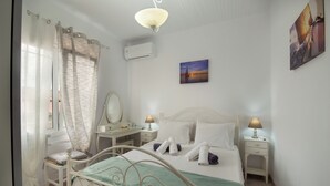 A bedroom with a bed, bedside tables, a desk, a chair, a window with curtains, an air conditioner, and a painting on the wall.