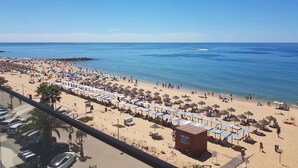 Een strand met rijen parasols en ligbedden, een klein gebouw met de tekst 'pista. Vidol' erop, en een helderblauwe lucht.