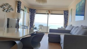 Een moderne woonkamer met een bank, een eethoek met een tafel en stoelen, en uitzicht op zee door grote ramen.