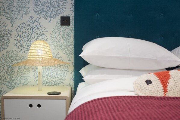Un lit avec des oreillers blancs et une couverture au point de crochet, une table de chevet avec une lampe, et un mur orné d’un motif corail.