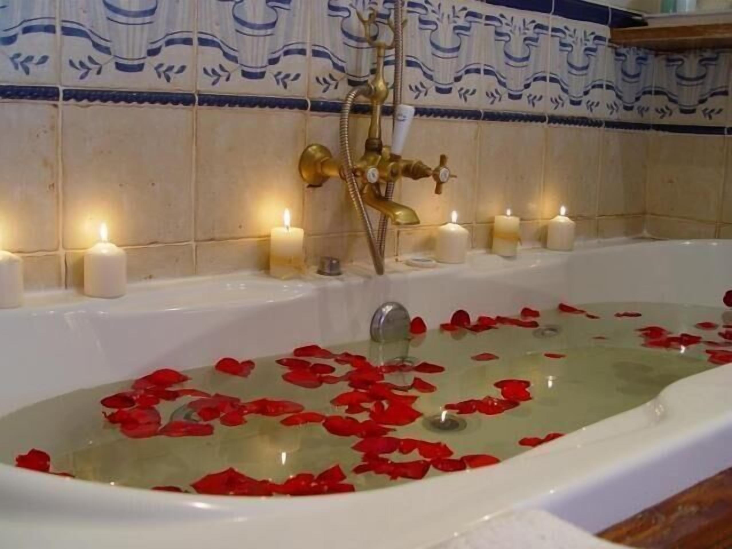 A bathtub filled with water and rose petals, surrounded by candles and a gold faucet.