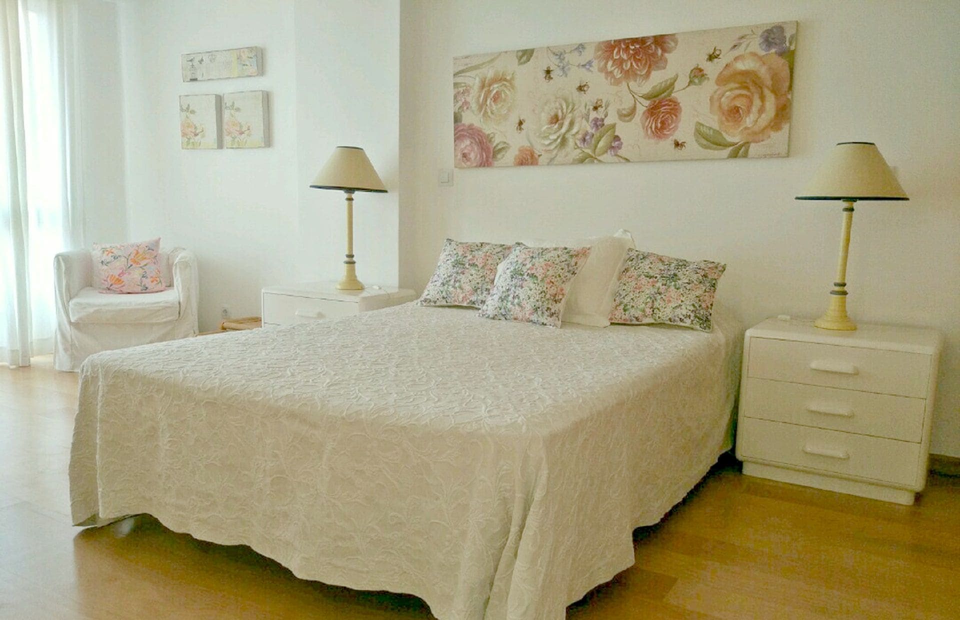 A bedroom with a large bed, two bedside tables with lamps, a chair, and a floral painting on the wall.