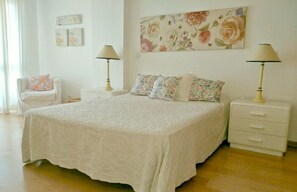 Une chambre à coucher avec un grand lit, deux tables de chevet avec des lampes, une chaise et un tableau représentant des fleurs accroché au mur.
