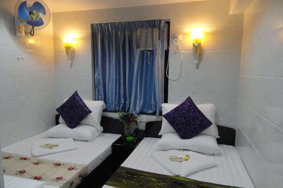 A hotel room with two beds, each with a pillow and a small table. There is a wall-mounted fan and two wall sconces with lights.