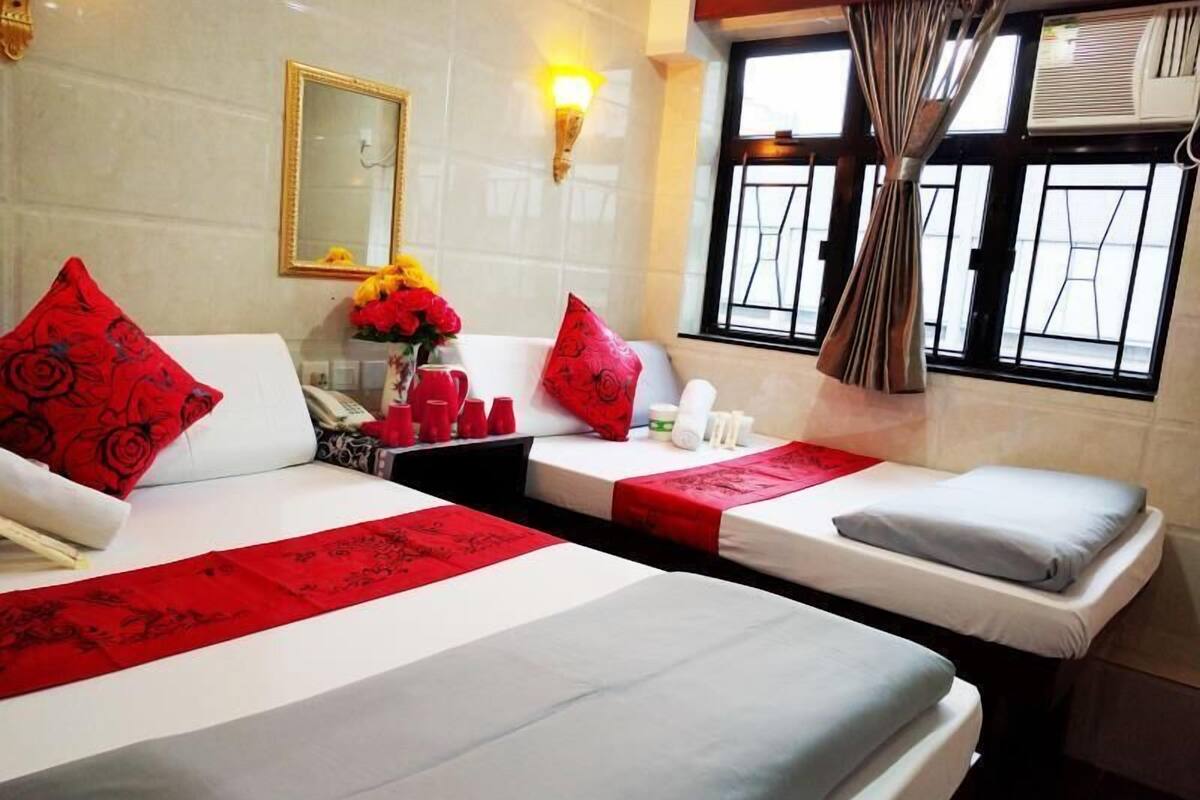 A hotel room with two beds, a bedside table with a telephone, a vase with flowers, and a window with curtains.