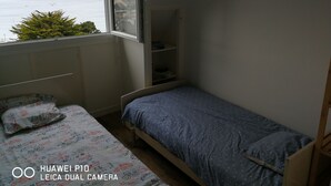 A bedroom with a bed, a window with a view of the sea, and a small shelf unit.