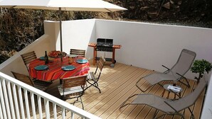 A wooden deck with a table set for dining, a grill, and two lounge chairs.