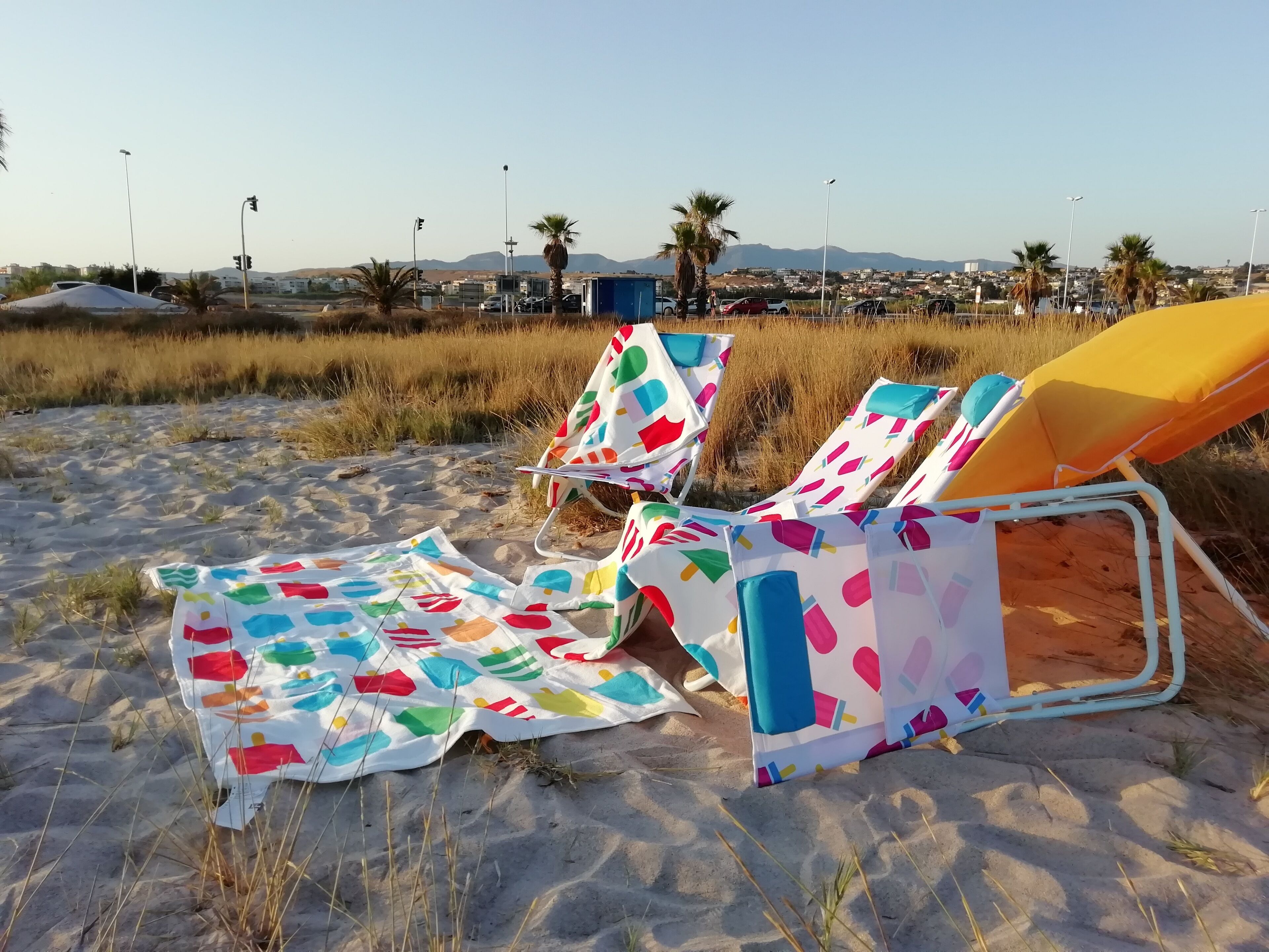 Sillas de playa con fundas coloridas en una playa arenosa, con palmeras y un paisaje urbano al fondo.