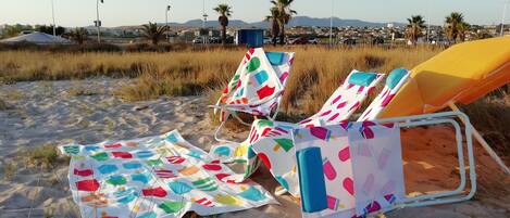 Sillas de playa con fundas coloridas en una playa arenosa, con palmeras y un paisaje urbano al fondo.