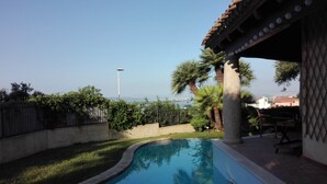 Een zwembad met een gebogen rand, omgeven door gazon en palmbomen. Er is een pergola met een betegeld dak en een tafel en stoelen eronder.