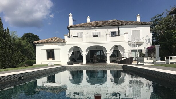 En stor, hvid bygning med tegltag, swimmingpool og træterrasse.