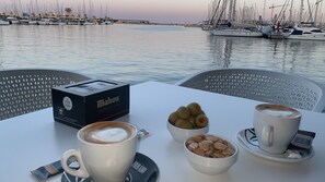 A table with a box of Mahou coffee, two cups of coffee, a bowl of almonds, and a bowl of pastries at a waterfront cafe. - Boutique Apartment by the Sea (Alicante)