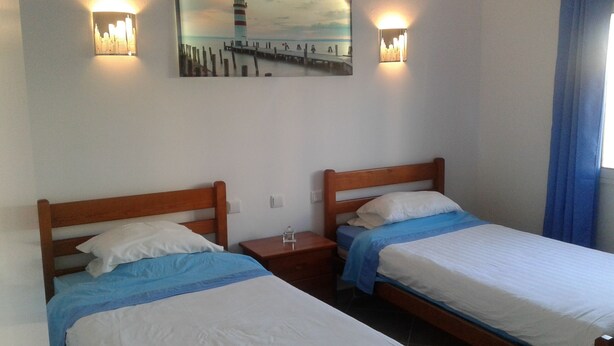 Una habitación con dos camas de madera, cada una con una almohada y una manta azul. Hay luces fijadas en la pared sobre las camas y un cuadro de un faro en la pared.