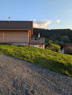 Un edificio in legno con un balcone, circondato da un vialetto ghiaioso e prato, con una collina boscosa sullo sfondo.