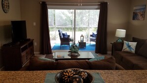Una sala de estar con televisión, ventilador de techo, una mesa de comedor con un cuenco de adornos y vistas a un patio con mobiliario exterior.
