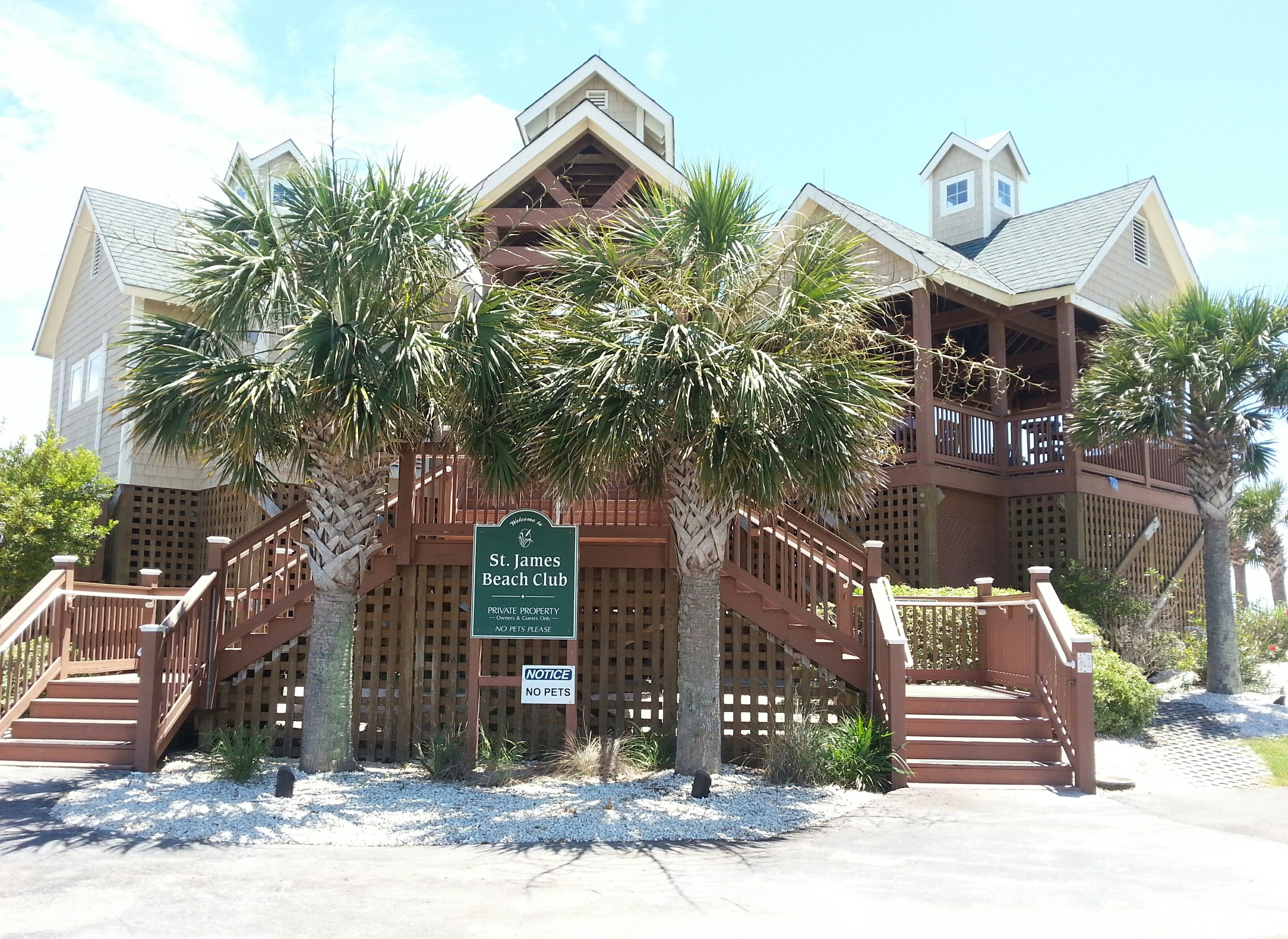 A two-story building with a wooden deck and stairs, surrounded by palm trees. A sign indicates it is the St. James Beach Club, private property, and no pets are allowed.