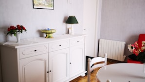 Un comedor con una mesa y sillas blancas, un armario blanco con cajones, una lámpara verde y un cuadro en la pared.