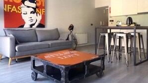 Een moderne woonkamer met een grijze bank, een salontafel, een keukenhoek met een barkruk, en een grote poster aan de muur.