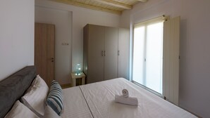 A bedroom with a bed, pillows, a bedside table with a lamp, a wardrobe, and a window with blinds. - Cycladic Breeze Villa Aura with swimming pool villa & great sea view  (Paros)
