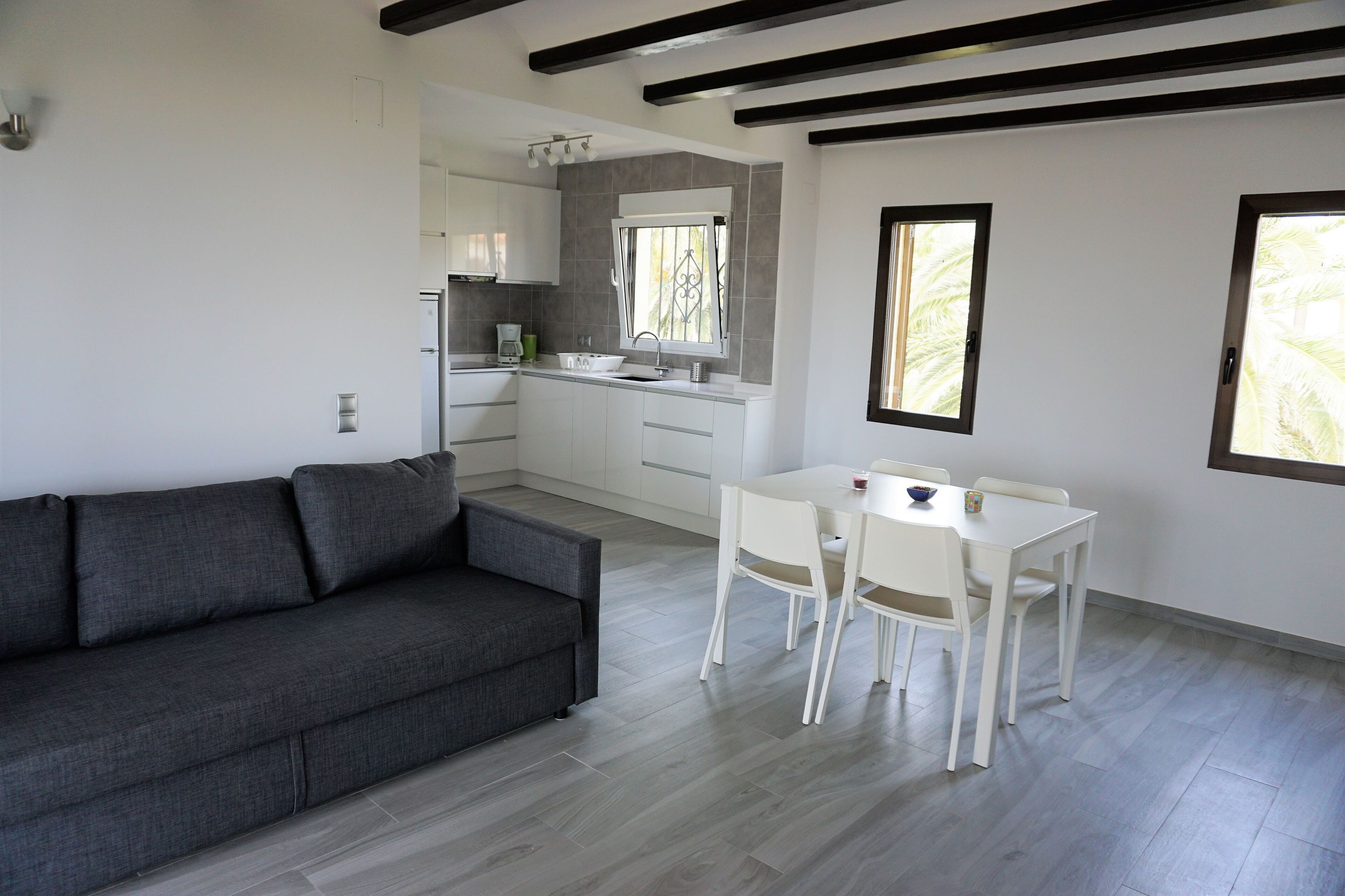Eine moderne Küche mit weißen Schränken und einer grauen Arbeitsplatte, ein Essbereich mit einem weißen Tisch und Stühlen sowie ein graues Sofa im Wohnbereich.