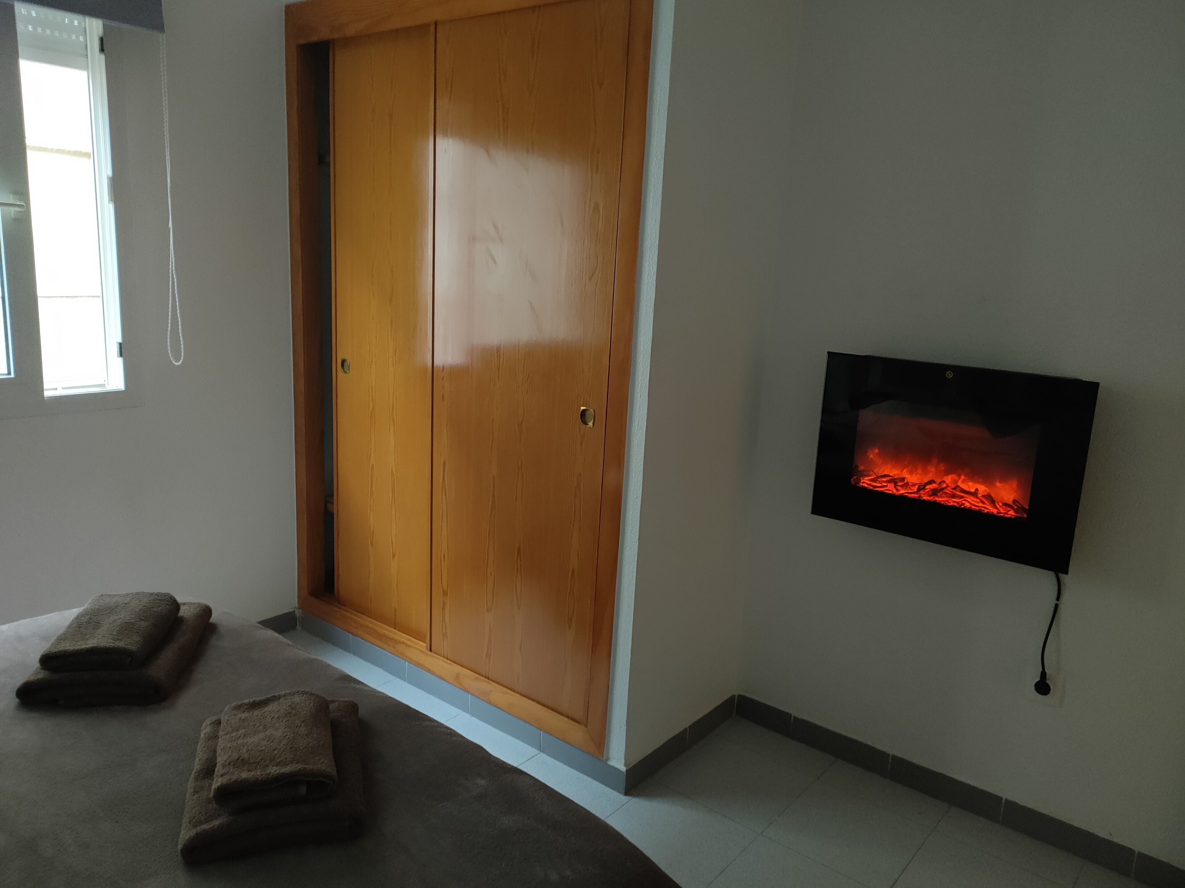 A room with a wooden wardrobe, a flat-screen TV displaying a fireplace, and two towels on the floor.