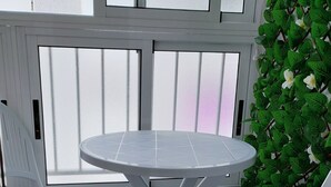 En liten utomhusplats med två vita plaststolar och ett runt bord, omgiven av en vit vägg med ett fönster och en grön växt med vita blommor.
