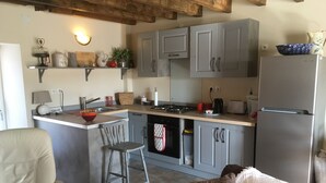 A compact kitchen with grey cabinets, a central island with a sink, a stove, and a refrigerator. There is a bar stool and a cushioned chair in the seating area.