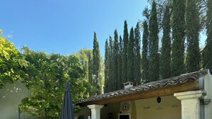 A patio area with three wicker lounge chairs, a table, and an umbrella. There is a small seating area with a sofa and a coffee table. The patio is surrounded by a low wall and has a view of tall trees. - Exceptional villa in Provence's Luberon region with a pool and breathtaking views (CAVAILLON)
