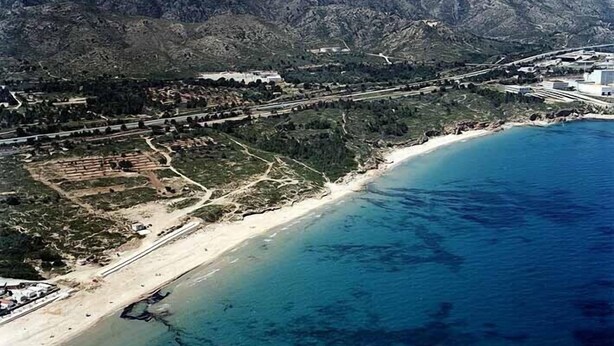 Vue aérienne d’une zone côtière avec une plage, une route et un paysage montagneux en arrière-plan.