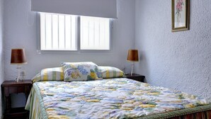 Une chambre à coucher avec un lit, deux tables de chevet, une fenêtre et un cadre photo fixé au mur.