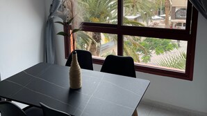 A dining area with a table and chairs, a vase with dried plants, and a view of palm trees and buildings through a window. - Casa Jandía is an apartment with terrace in the center and 100 meters from the beach. (Morro Jable)