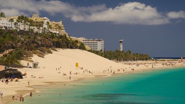 A beach with clear turquoise waters, a sandy shore, and a building with a lighthouse in the background. - Casa Jandía is an apartment with terrace in the center and 100 meters from the beach. (Morro Jable)