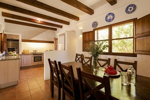 A kitchen with wooden beams, a dining table with chairs, and a window with a view of greenery.
