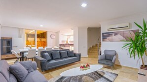 A modern living room with a grey sofa, a coffee table with flowers, a dining area with a table and chairs, and a staircase leading to another level.
