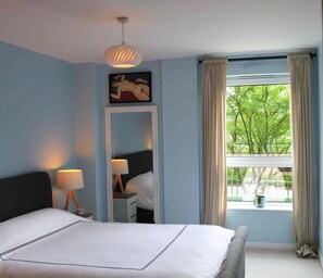 A bedroom with a bed, bedside lamps, a nightstand, a dresser, a mirror, a window with curtains, and a ceiling light fixture.